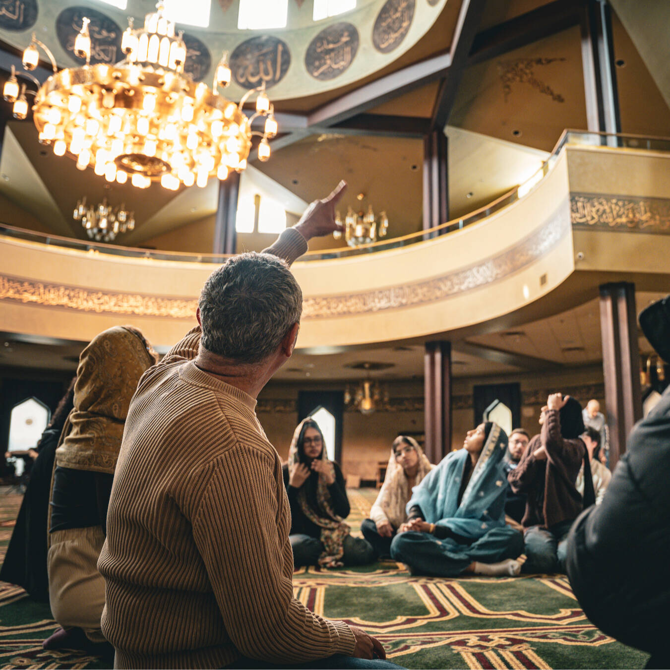 Over the shoulder of a gentleman pointing to the second floor of a masjid prayer space with a large chandelier.